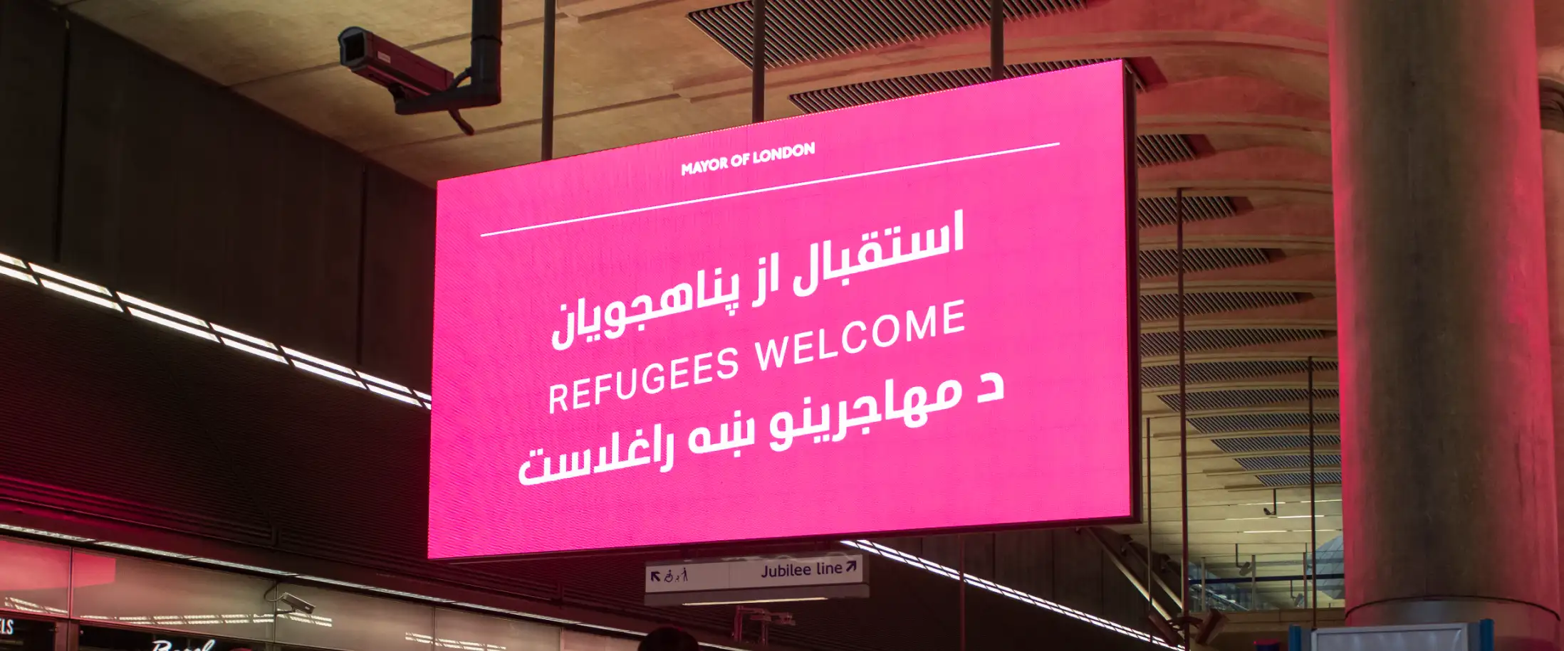 A bright pink LED display advert in a London Underground station with text in Dari, English and Pashto reading 'Refugees Welcome'